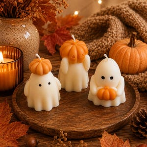 Op de afbeelding: Drie gebroken witte, spookvormige kaarsen met zwarte stippen als ogen, elk met een kleine oranje pompoen. Eén spook draagt een pompoen als hoed, een ander heeft een pompoen op het hoofd en de derde houdt een kleine oranje pompoen in de handen.