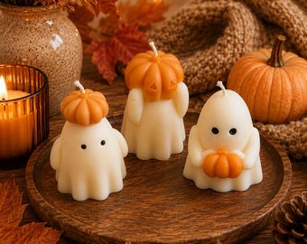 May include: Three off-white ghost-shaped candles with black dot eyes, each featuring a small orange pumpkin. One ghost wears a pumpkin as a hat, another has a pumpkin on its head, and the third holds a small orange pumpkin in its hands.
