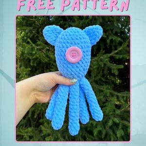 Puede incluir: Un pulpo de ganchillo azul hecho a mano con un ojo de botón rosa y orejas caídas. El texto "FREE PATTERN" se muestra en la parte superior de la imagen. El pulpo se sostiene en una mano sobre un fondo verde.