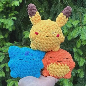 May include: Three crocheted plush toys in the shape of popular characters. The top toy is yellow with brown ears and red cheeks. Below it are a blue and an orange toy. All have closed eyes, suggesting they are sleeping.