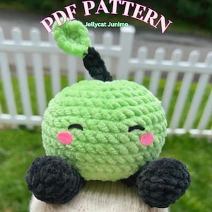 Puede incluir: Un juguete de peluche verde tejido a ganchillo con una cara sonriente, mejillas rosas y ojos negros. Tiene brazos negros y un tallo verde. El texto "PDF PATTERN" y "Jellycat Junimo" son visibles.