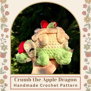 May include: A plush crochet dragon toy featuring a light green body, a tan shell resembling an apple pie crust, and red apple-like accents on its head and back. The text Crumb the Apple Dragon Handmade Crochet Pattern is displayed below the toy.