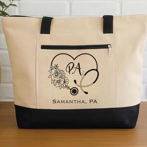 May include: A beige canvas tote bag with a black bottom panel and black shoulder straps. The front features a black graphic of a heart-shaped stethoscope intertwined with flowers, the letters PA, and the text SAMANTHA, PA printed below.