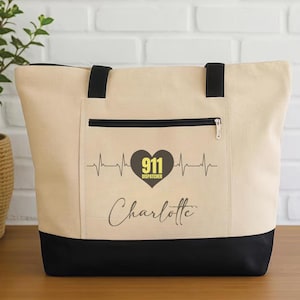 Könnte beinhalten: Beige Tragetasche mit schwarzen Akzenten, einschließlich einer Reißverschlusstasche vorne. Das Design zeigt ein herzförmiges Grafik mit "911 Dispatcher" und einem Namen, sowie eine Herzschlaglinie. Die Tasche hat schwarze Griffe.