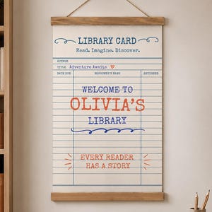 May include: A vertical wall hanging featuring a cream-coloured canvas printed with a library card design. The text reads: LIBRARY CARD, Read. Imagine. Discover., Adventure Awaits, WELCOME TO OLIVIA'S LIBRARY, and EVERY READER HAS A STORY. It has wooden rails.