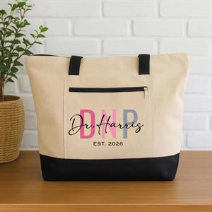 Può includere: Una borsa tote in tela beige con dettagli neri. Presenta una tasca con cerniera nera e il testo "DNP Dr. Harris EST. 2026" in rosa, grigio e nero. La base della borsa è nera, con manici neri.