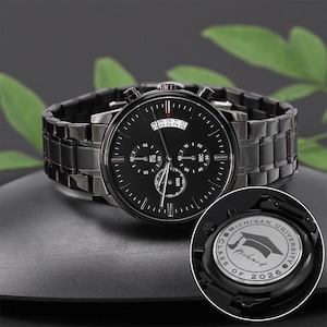 May include: A black wristwatch with a matching band. The watch face has multiple sub-dials and a date display. The back of the watch is engraved with "MICHIGAN UNIVERSITY", a graduation cap, and "CLASS OF 2026".