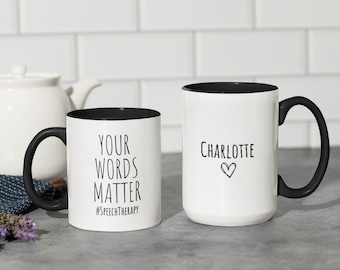 Op de afbeelding: Twee witte keramische mokken met zwarte binnenkant en handvatten. Eén mok leest "YOUR WORDS MATTER #SPEECHTHERAPY" in zwarte tekst. De andere mok heeft de naam "Charlotte" en een hartsymbool. Beide mokken staan op een grijze ondergrond.