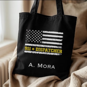 Könnte beinhalten: Schwarze Tragetasche mit einem verblassten amerikanischen Flaggen-Design und dem Text "911 DISPATCHER" in Gelb und Weiß. Der Name "A. MORA" ist darunter aufgedruckt. Die Tasche hat schwarze Griffe.