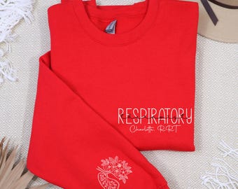 Może przedstawiać: Jasnoczerwona bluza z napisem "RESPIRATORY" w białym skrypcie na piersi. Rękaw zdobi biały kwiatowy i płucny wzór. Pod słowem "RESPIRATORY" znajduje się napis "Charlotte, RRT".