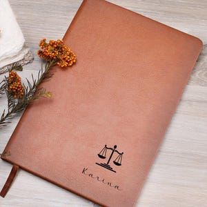 May include: A brown leather-bound journal with a debossed scales of justice symbol and the name "Karina" in script. The journal has a ribbon bookmark and is decorated with orange flowers.