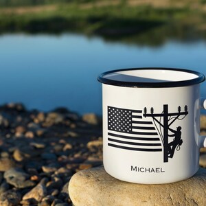 May include: White enamel mug with a black rim and handle. The mug features a black and white American flag design and a silhouette of a lineman on a utility pole. The name "MICHAEL" is printed below the design.