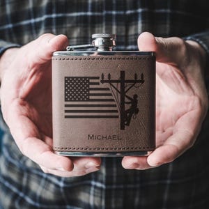 May include: A brown leather flask with a silver cap. The front features an engraved American flag, a utility pole with a lineman, and the name "MICHAEL". The flask is held in both hands.