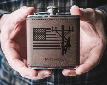 May include: A brown leather flask with a silver cap. The front features an engraved American flag, a utility pole with a lineman, and the name "MICHAEL". The flask is held in both hands.