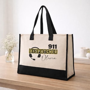 May include: A canvas tote bag with black handles and a black bottom panel. The bag features the text "911 DISPATCHER Olivia" in black and yellow, along with a headset graphic. The bag is sitting on a wooden surface.