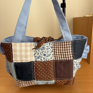 May include: A patchwork tote bag with a variety of fabric patterns in brown, blue, and cream. The bag features a light blue lining and handles, with a brown polka dot bow. The bag is approximately 10 inches wide.