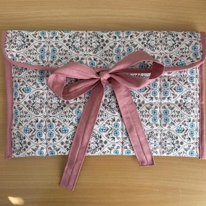 May include: A rectangular fabric case with a floral pattern in white, blue, and pink. The case is trimmed with pink fabric and tied with a pink ribbon bow. The case is likely used for storing items.