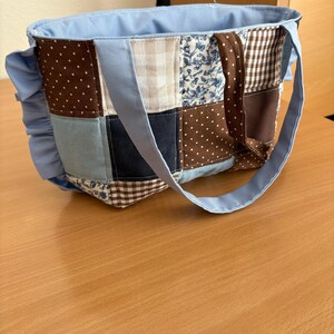 May include: A patchwork tote bag with a light blue lining and handles. The bag's exterior features a variety of fabric patterns, including polka dots, floral prints, and gingham checks in shades of blue, brown, and cream. A ruffled accent adorns one side.