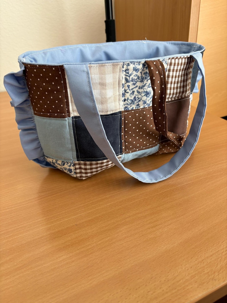 May include: A patchwork tote bag with a light blue lining and handles. The bag's exterior features a variety of fabric patterns, including polka dots, floral prints, and gingham checks in shades of blue, brown, and cream. A ruffled accent adorns one side.