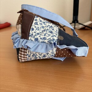 May include: A handmade patchwork bag with a light blue handle and ruffled trim. The bag features various fabric patterns, including floral, polka dot, and gingham, in shades of blue, brown, and white. The bag is designed with a unique shape and a light blue trim.
