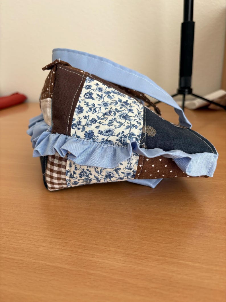May include: A handmade patchwork bag with a light blue handle and ruffled trim. The bag features various fabric patterns, including floral, polka dot, and gingham, in shades of blue, brown, and white. The bag is designed with a unique shape and a light blue trim.