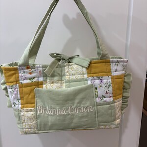May include: A handmade patchwork tote bag with a light green and yellow color scheme. The bag features a floral pattern, a front pocket with embroidered text, and light green handles. The bag is held up by a hand.