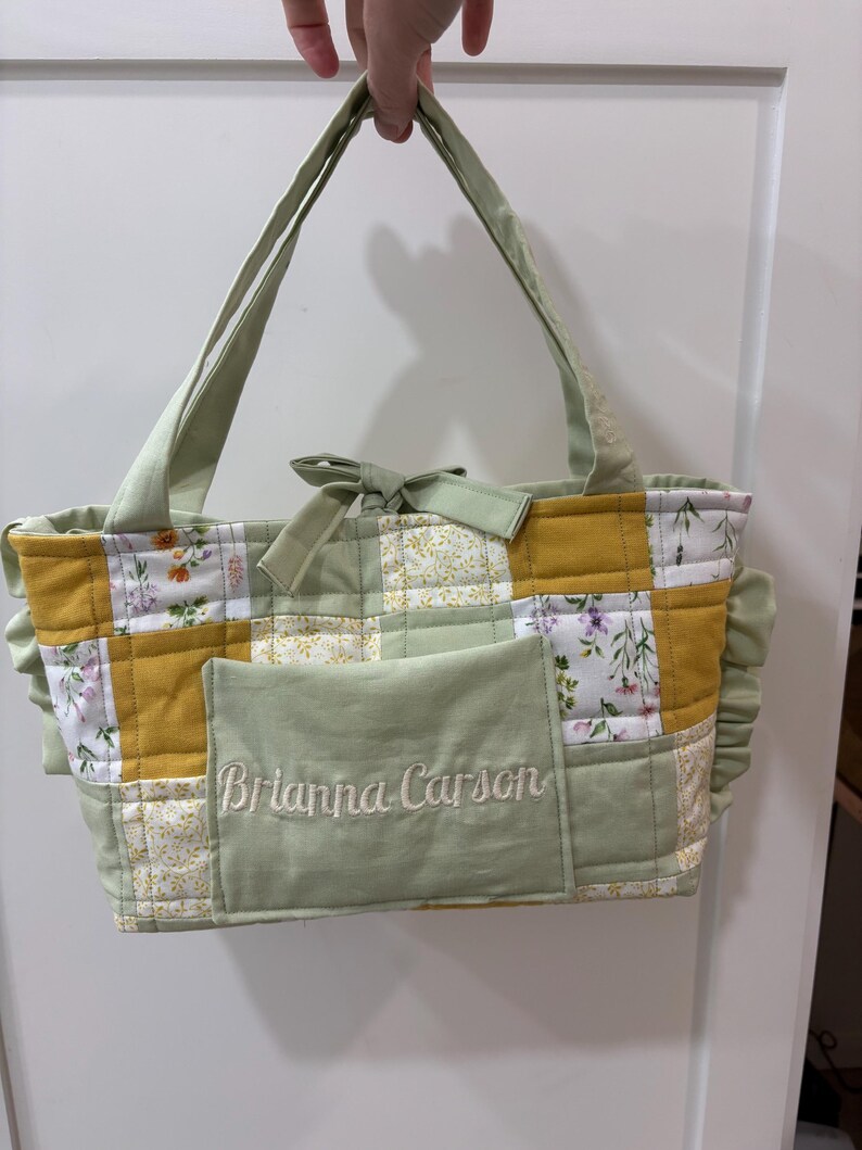 May include: A handmade patchwork tote bag with a light green and yellow color scheme. The bag features a floral pattern, a front pocket with embroidered text, and light green handles. The bag is held up by a hand.