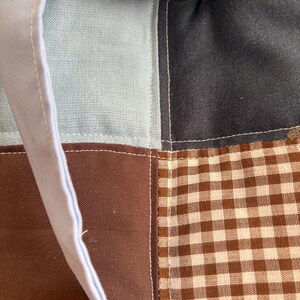 May include: Close-up of a patchwork tote bag with a light blue handle and a brown polka dot bow. The bag features squares of light blue, dark blue, brown, and brown and white gingham fabric, all stitched together with white thread.