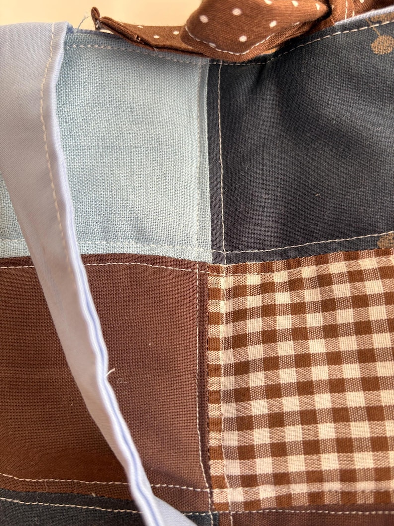May include: Close-up of a patchwork tote bag with a light blue handle and a brown polka dot bow. The bag features squares of light blue, dark blue, brown, and brown and white gingham fabric, all stitched together with white thread.