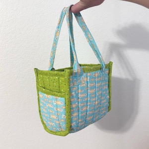 May include: A small, quilted tote bag with a fish pattern. The bag features a light blue background with yellow and orange fish, green polka dot accents, and matching handles. The bag is held up by a hand.