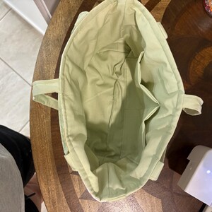May include: A light green tote bag with two handles and interior pockets. The bag is made of a soft fabric and appears to be handmade. The interior and exterior are the same color. The bag is sitting on a wooden table.