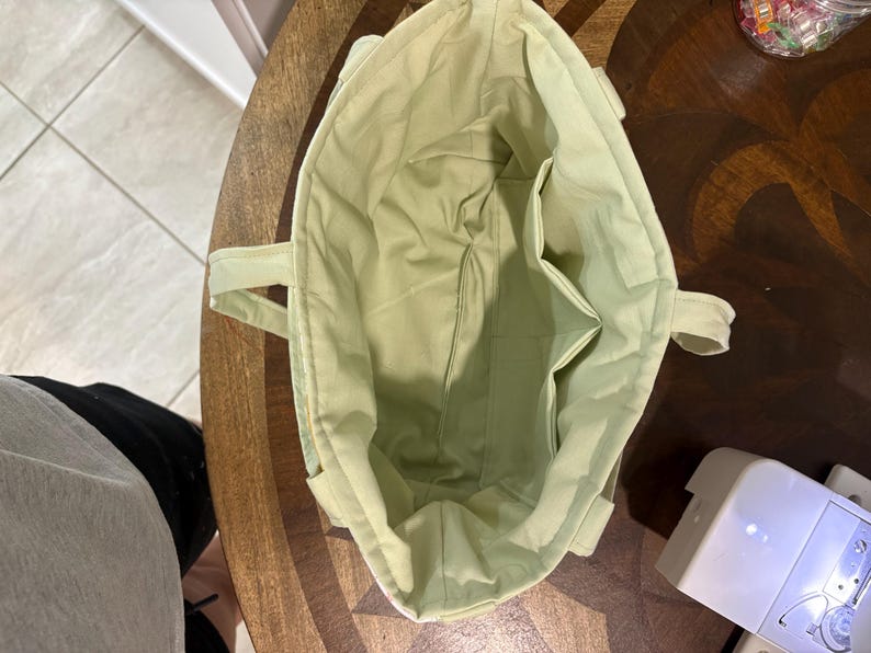 May include: A light green tote bag with two handles and interior pockets. The bag is made of a soft fabric and appears to be handmade. The interior and exterior are the same color. The bag is sitting on a wooden table.