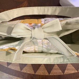 May include: A light green tote bag with a bow closure. The bag features a floral patterned interior and a light green handle. The bag is made of a soft fabric and has a rectangular shape.