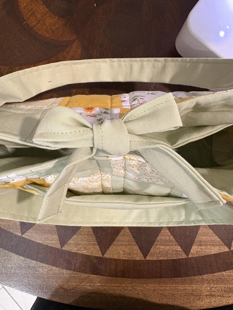 May include: A light green tote bag with a bow closure. The bag features a floral patterned interior and a light green handle. The bag is made of a soft fabric and has a rectangular shape.