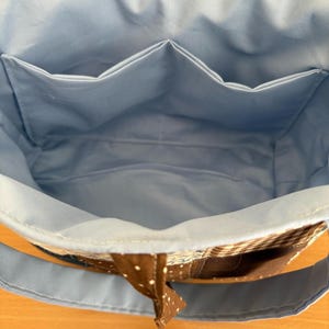 May include: A light blue tote bag with a brown polka dot handle and a light blue interior. The bag has three interior pockets. The exterior features a brown and white checkered pattern.