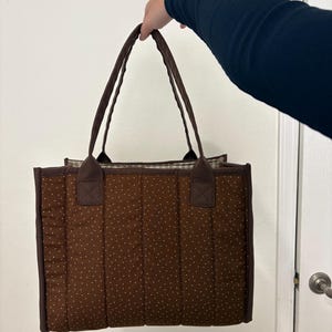 May include: A brown tote bag with white polka dots. The bag has two brown handles and a rectangular shape. The bag is made of quilted fabric and has a solid brown border.