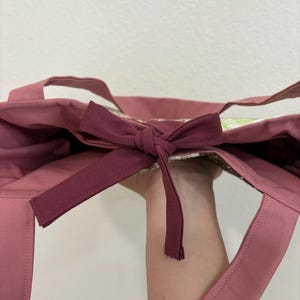 May include: A dusty rose tote bag with matching handles and a bow closure. The bag's interior features a floral pattern. The bow is a darker shade of rose. The bag is made of a sturdy fabric.