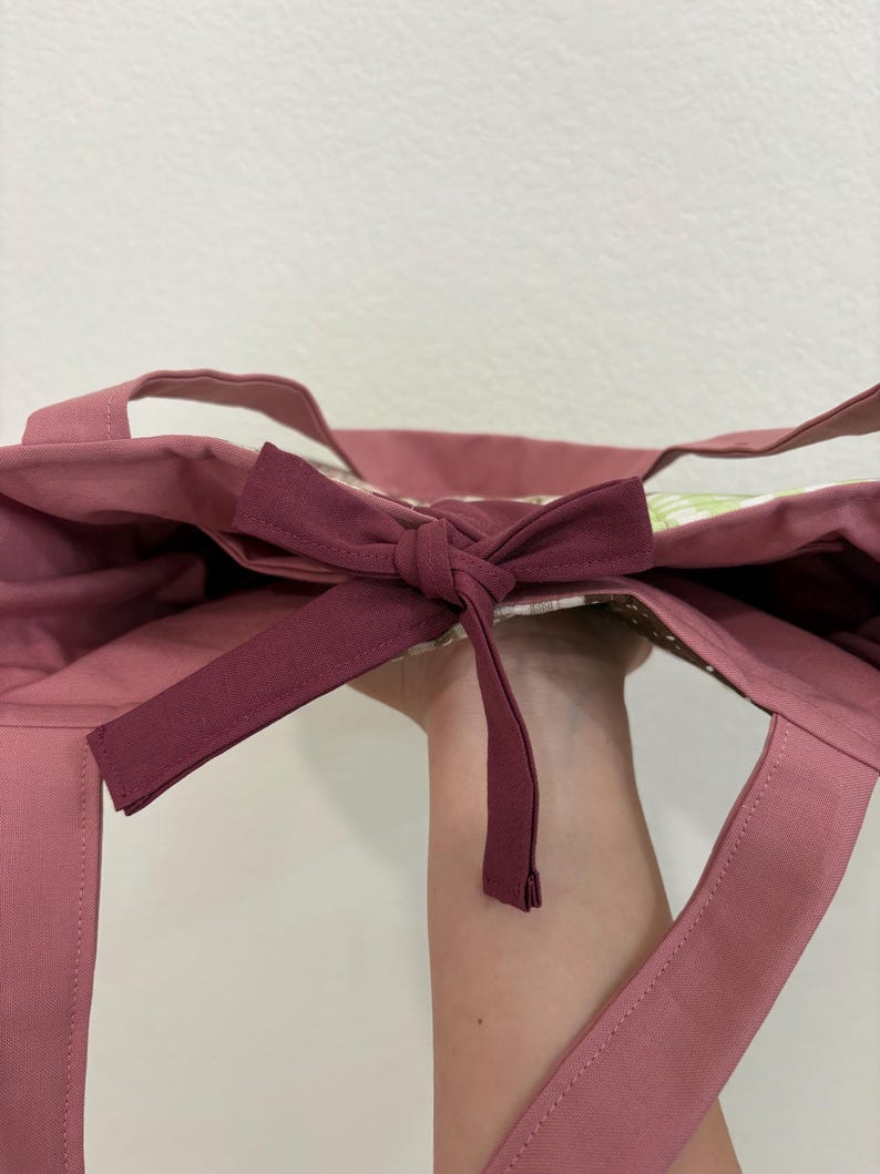 May include: A dusty rose tote bag with matching handles and a bow closure. The bag's interior features a floral pattern. The bow is a darker shade of rose. The bag is made of a sturdy fabric.