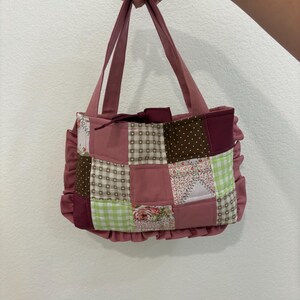 May include: A patchwork tote bag in shades of pink, burgundy, and brown. The bag features a variety of patterned fabric squares, including floral, polka dot, and gingham designs. The bag has a ruffled trim and matching pink handles.