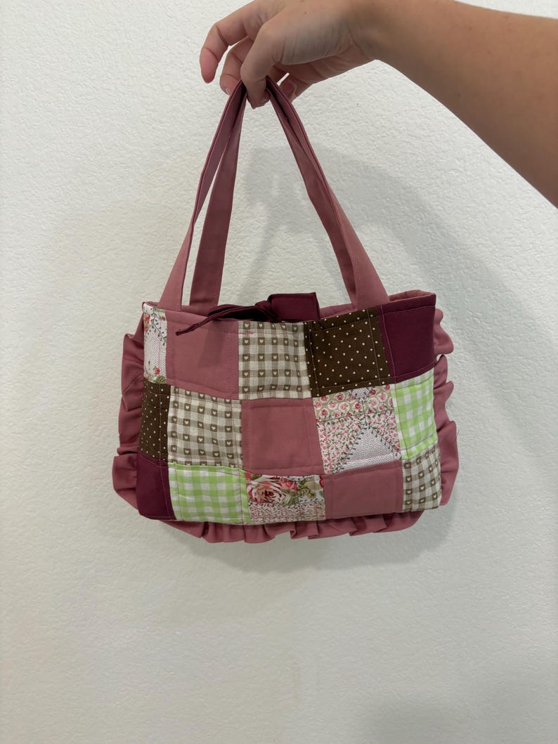 May include: A patchwork tote bag in shades of pink, burgundy, and brown. The bag features a variety of patterned fabric squares, including floral, polka dot, and gingham designs. The bag has a ruffled trim and matching pink handles.