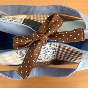 May include: A light blue tote bag with a brown polka dot bow. The bag features a patchwork design with various patterns, including floral and gingham, in shades of blue, brown, and white. The handles are a matching light blue.