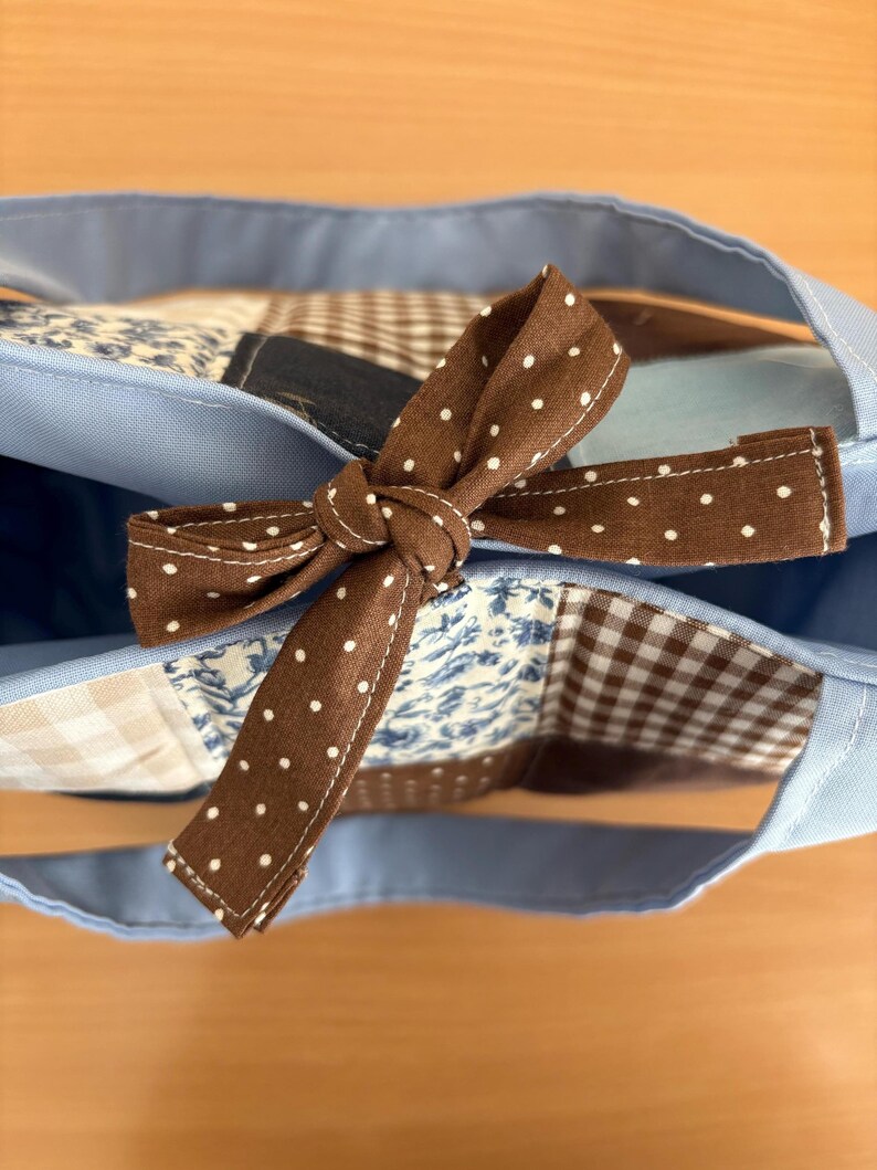 May include: A light blue tote bag with a brown polka dot bow. The bag features a patchwork design with various patterns, including floral and gingham, in shades of blue, brown, and white. The handles are a matching light blue.