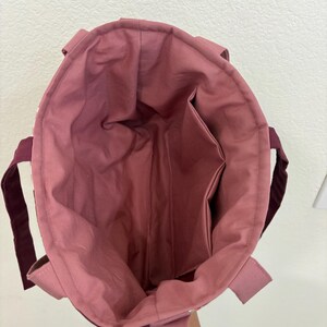 May include: Open view of a tote bag with a mauve interior and exterior. The bag has a rectangular shape with a deep interior pocket. The handles are a dark burgundy color.