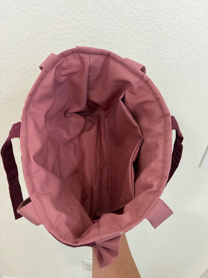 May include: Open view of a tote bag with a mauve interior and exterior. The bag has a rectangular shape with a deep interior pocket. The handles are a dark burgundy color.