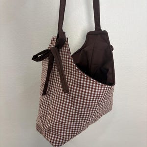 May include: A brown and white gingham tote bag with brown straps and a bow detail. The bag has a simple, open design and is made of fabric. The interior of the bag is a solid brown color.