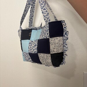 May include: A patchwork tote bag with a floral pattern and various shades of blue, black, and gray squares. The bag has matching floral handles and a ruffled edge.