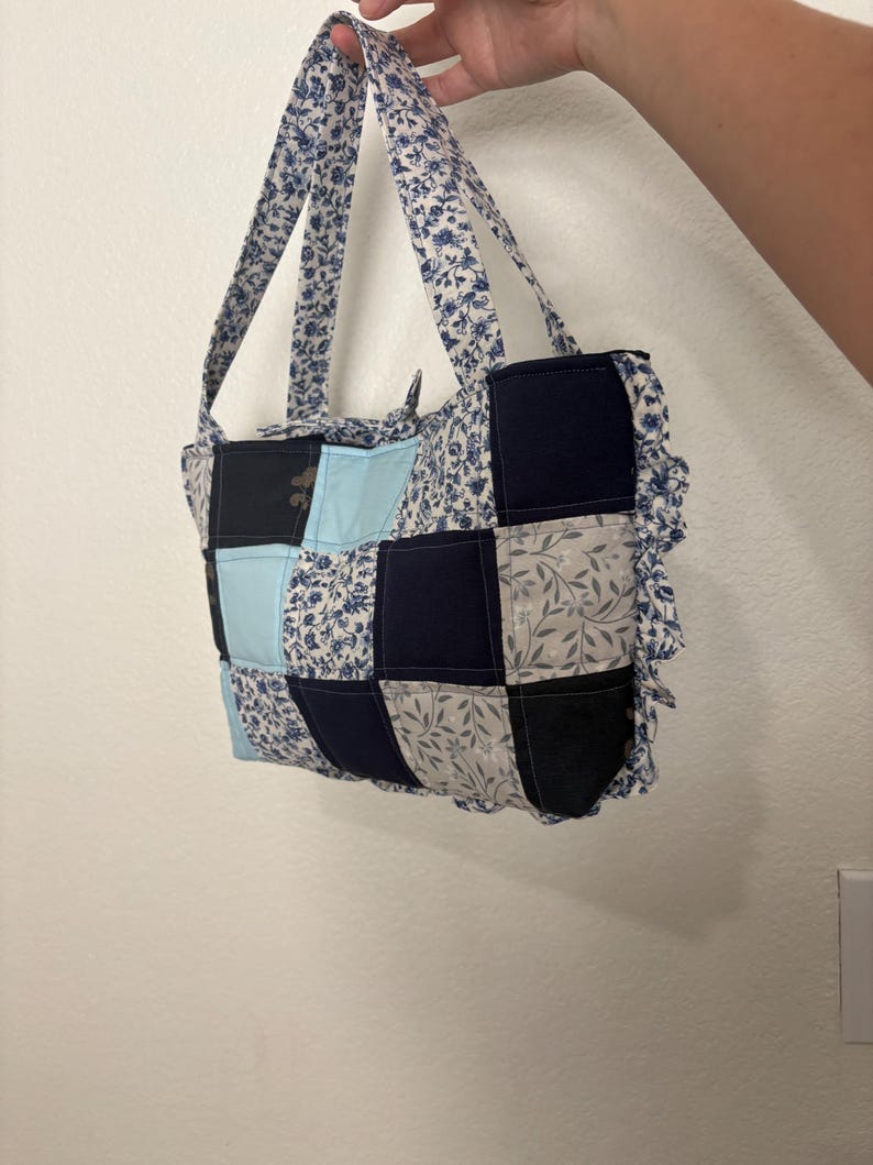 May include: A patchwork tote bag with a floral pattern and various shades of blue, black, and gray squares. The bag has matching floral handles and a ruffled edge.