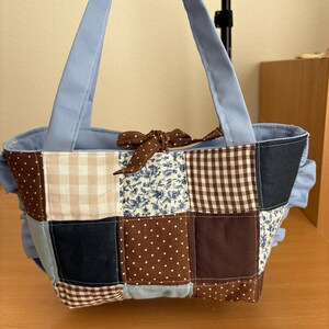 May include: A patchwork tote bag with a light blue lining and handles. The bag features a variety of fabric squares in shades of brown, blue, and cream, including polka dots, floral prints, and gingham patterns. A brown bow adorns the top.