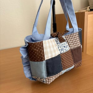 May include: A patchwork tote bag with a light blue interior and handles. The bag features a variety of fabric squares in brown, blue, and cream with polka dots, gingham, and floral patterns. A brown bow is attached to the top.