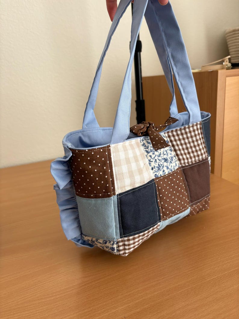 May include: A patchwork tote bag with a light blue interior and handles. The bag features a variety of fabric squares in brown, blue, and cream with polka dots, gingham, and floral patterns. A brown bow is attached to the top.